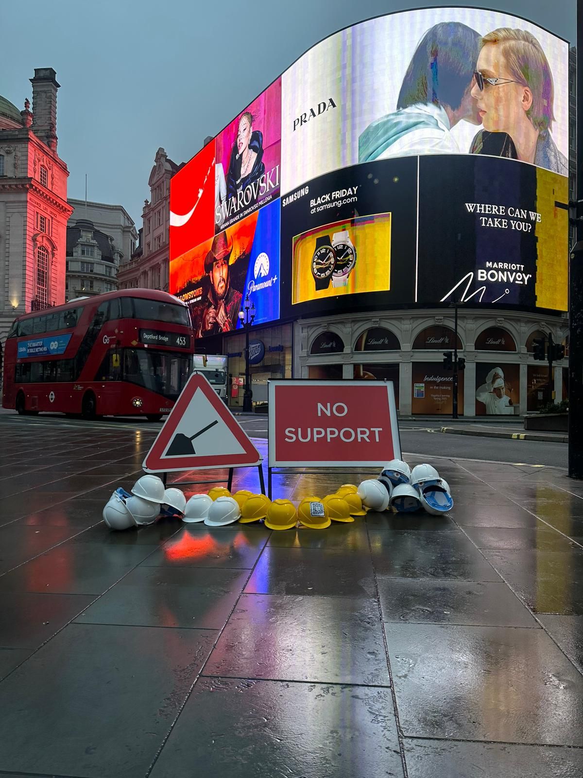 Two construction signs in London's Piccadilly Circus, one showing a spade and another one saying 'no support'. There are yellow and white hard hat helmets around them.