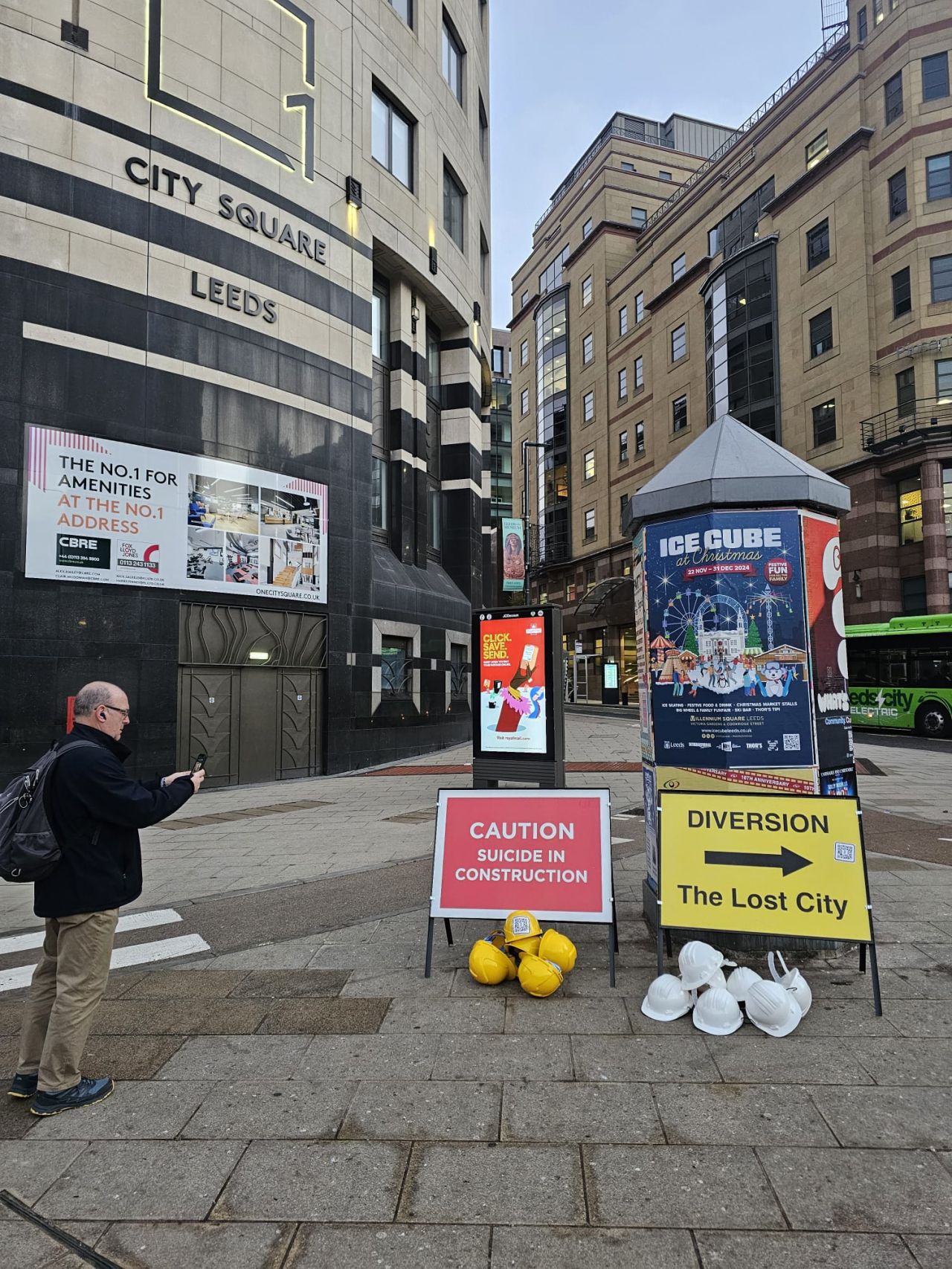 Two construction signs in Leeds' city square, one saying 'caution, suicide in construction' and the other 'diversion, the lost city' with an arrow pointing to the right. There white hard hat helmets around them.