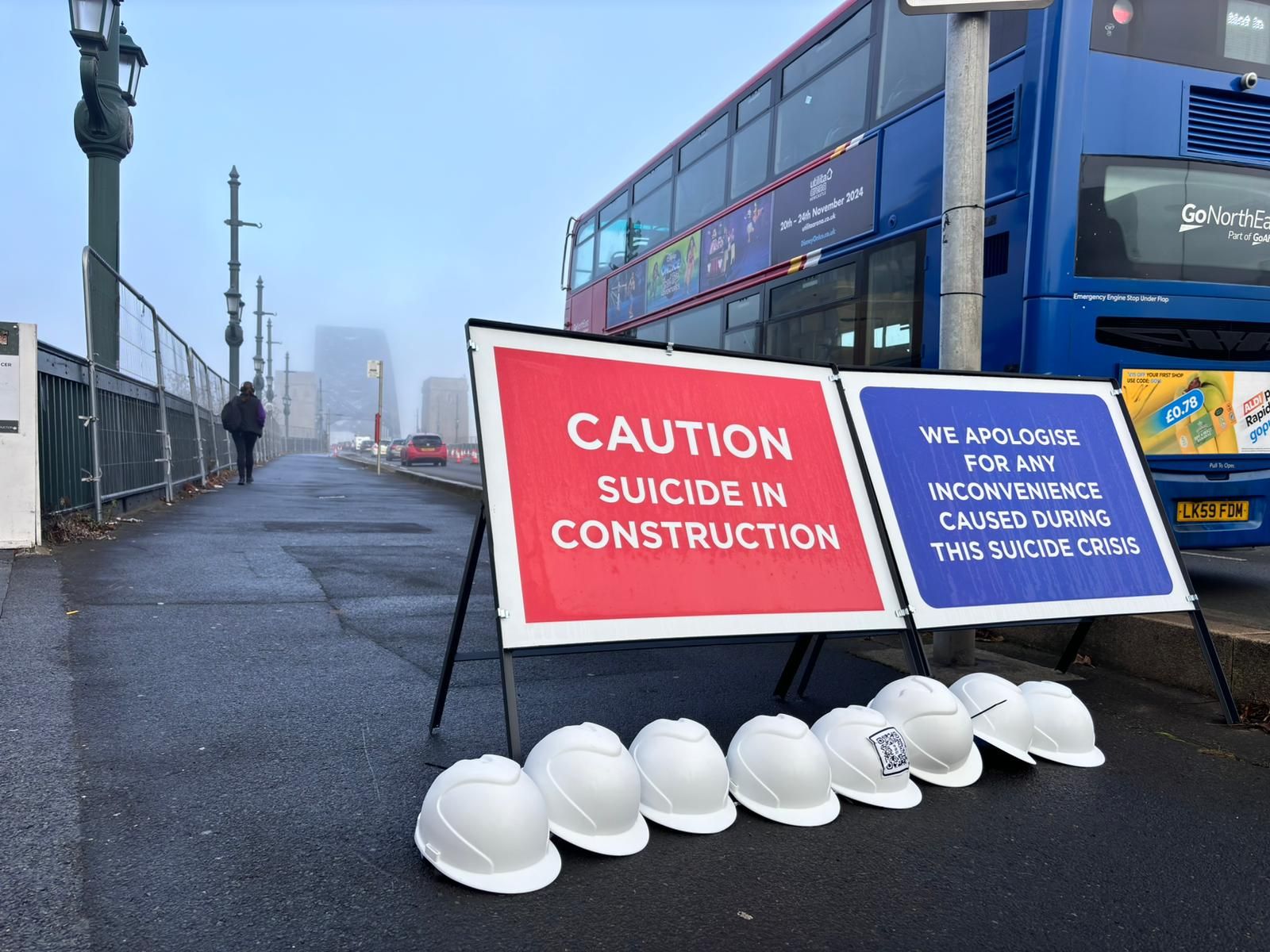 Two construction signs on a road with a bus on the background, one saying 'caution, suicide in construction' and the other 'we apologise for any inconvenience caused during this suicide crisis'. There white hard hat helmets around them.