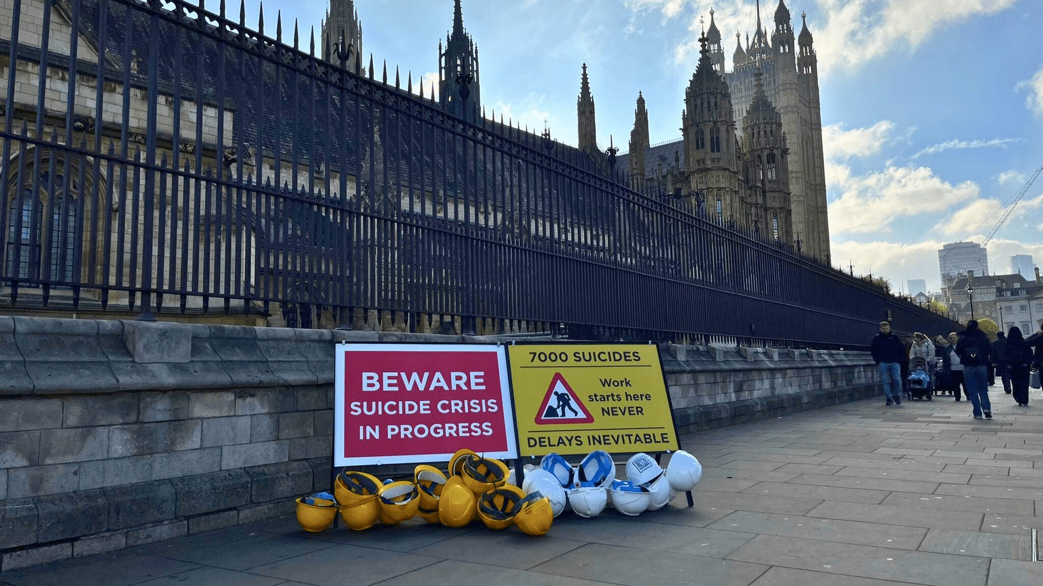 Two construction signs outside the UK Parliament that say 'beware, suicide crisis in progress' and '7,000 suicides. Work starts here NEVER. Delays inevitable'. There are yellow and white hard hat helmets around them.